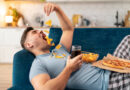 homem deitado no sofá comendo salgadinhos e tomando refrigerante representando hábitos que prejudicam