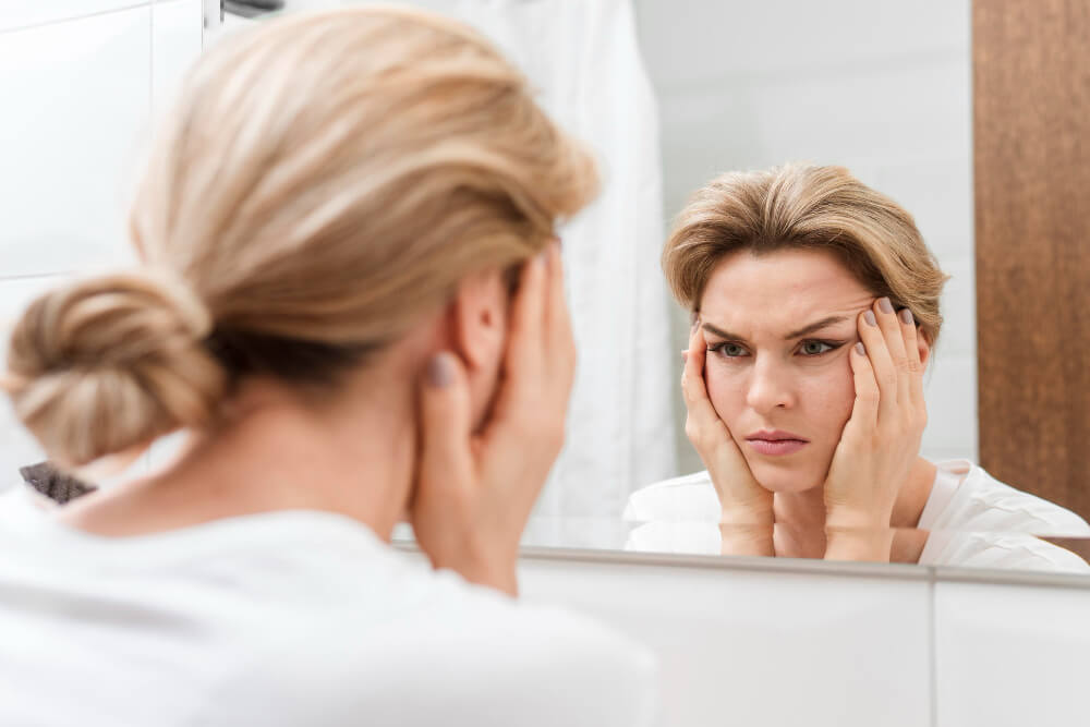 Mulher observa o rosto no espelho com expressão de preocupação, em referência à trend “cortisol face” e ao inchaço facial.