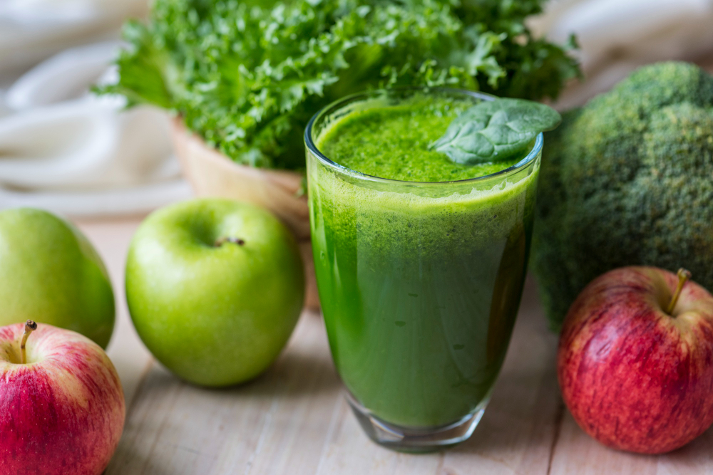 Copo de suco verde com maçãs, folhas e vegetais ao redor, representando a tendência de detox do fígado