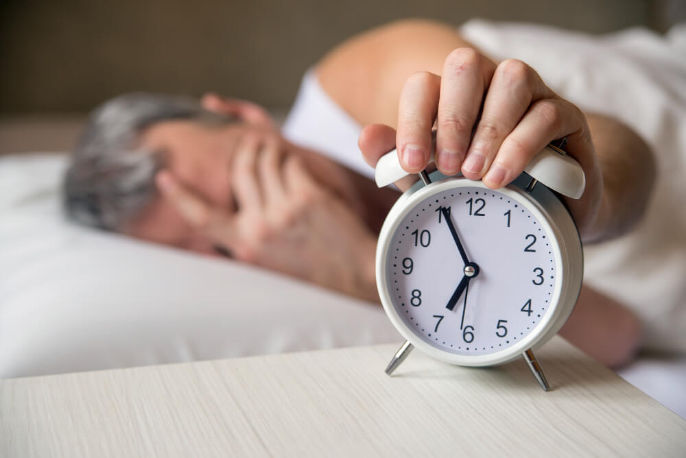 Pessoa acordando cansada e segurando despertador ao lado da cama, representando baixa qualidade do sono