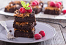 Brownie de chocolate com framboesas, hortelã e cobertura cremosa, servido em prato cinza sobre mesa de madeira