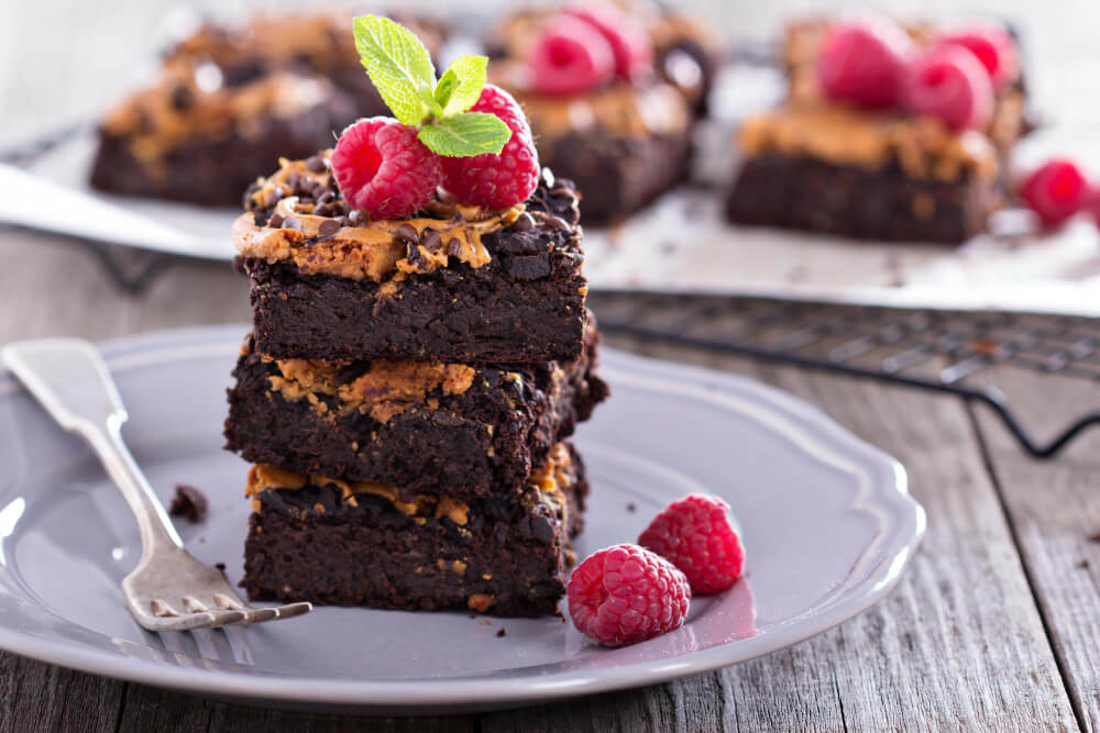 Brownie de chocolate com framboesas, hortelã e cobertura cremosa, servido em prato cinza sobre mesa de madeira