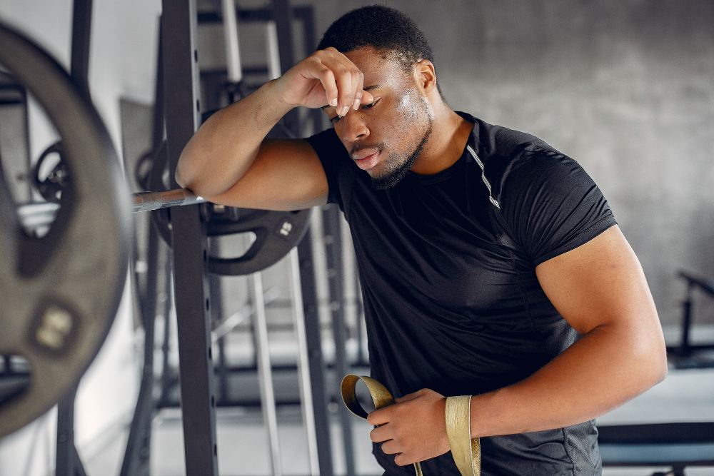 Homem exausto apoiado em barra de musculação durante treino de academia, com a mão na testa