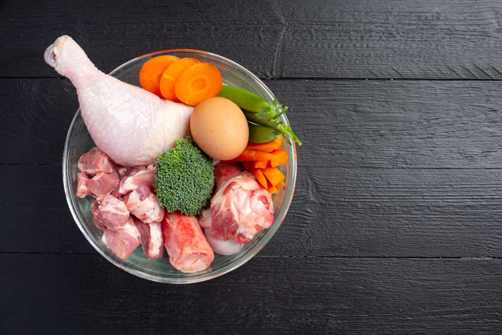Tigela de vidro com coxa de frango crua, cubos de carne, ovo, brócolis e cenoura, fontes de proteína sobre mesa de madeira escura