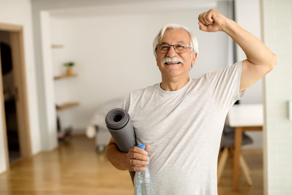 Homem idoso sorridente com o punho erguido, segurando tapete de exercício e garrafa de água, símbolo de vitalidade e healthspan