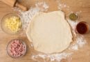 Massa de pizza tradicional sendo aberta com rolo, cercada de queijo ralado, presunto em cubos, orégano e molho de tomate