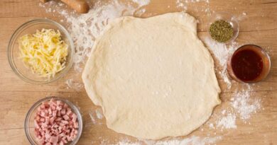 Pizza de massa proteica: a receita de queijo cottage que viralizou Massa de pizza tradicional sendo aberta com rolo, cercada de queijo ralado, presunto em cubos, orégano e molho de tomate