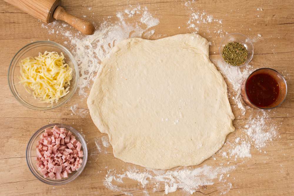 Massa de pizza tradicional sendo aberta com rolo, cercada de queijo ralado, presunto em cubos, orégano e molho de tomate
