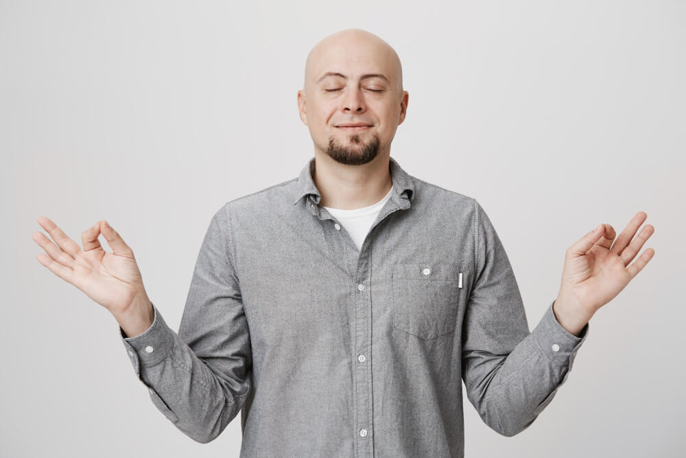 Homem calvo e barbudo, de olhos fechados e expressão serena, mãos em gesto de meditação