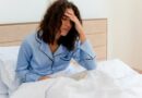 Mulher sentada na cama com a mão na testa, com expressão de quem acordou cansada
