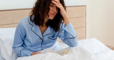 Você acorda cansado mesmo depois de dormir? Veja o que pode estar acontecendo Mulher sentada na cama com a mão na testa, com expressão de quem acordou cansada