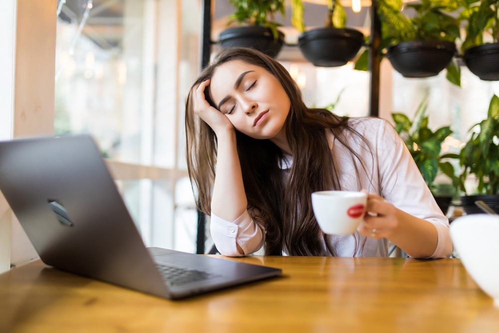 Mulher com cansaço e falta de energia sentada à mesa com notebook e xícara de café