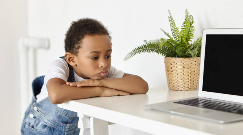 Criança cansada apoiada nos braços em frente a um notebook, em mesa de escritório doméstico