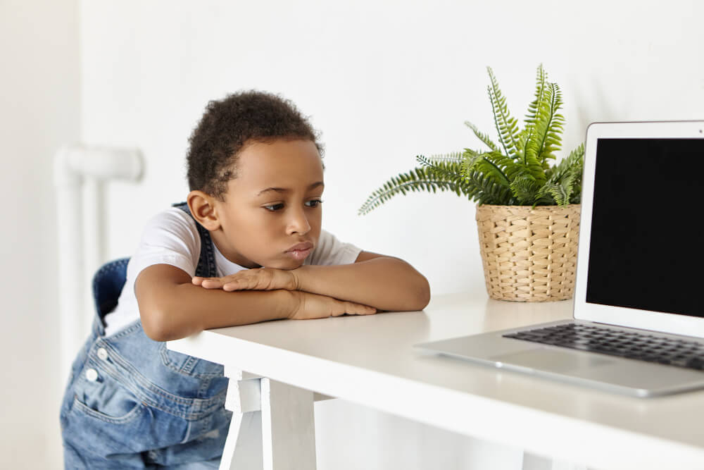 Criança cansada apoiada nos braços em frente a um notebook, em mesa de escritório doméstico