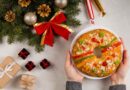 Bolo de Natal em formato de coroa com frutas cristalizadas e amêndoas, servido em prato branco entre enfeites natalinos