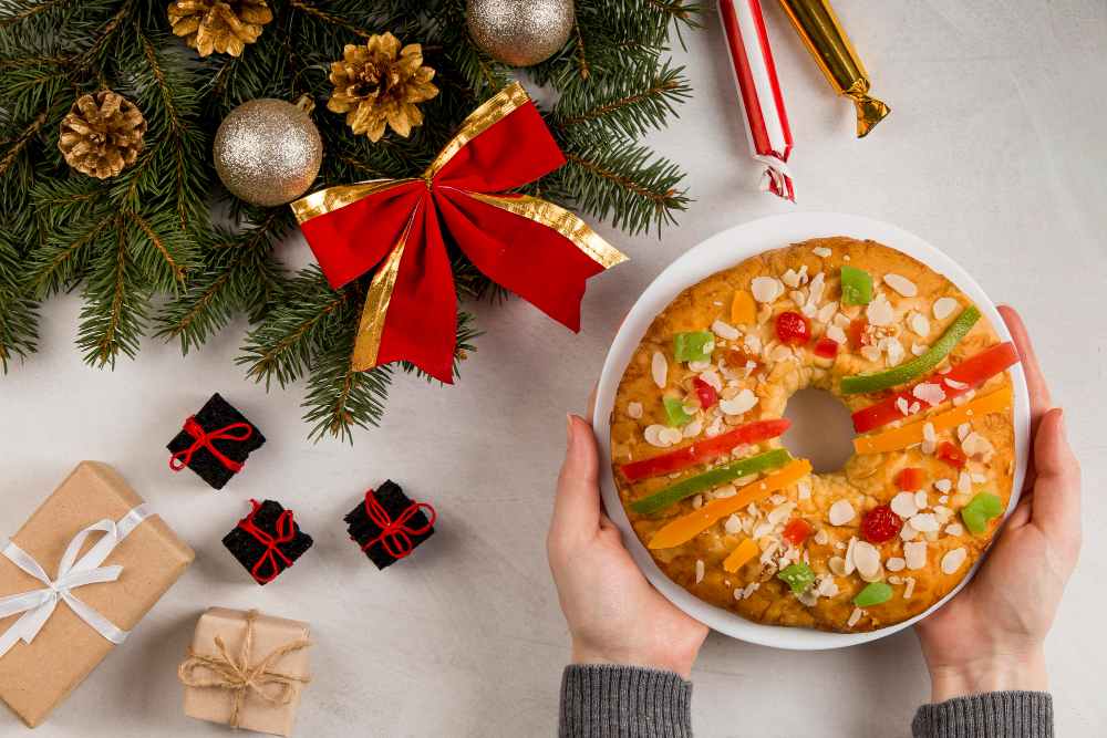 Bolo de Natal em formato de coroa com frutas cristalizadas e amêndoas, servido em prato branco entre enfeites natalinos