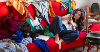 Organização da casa: a bagunça afeta sua rotina? Teste em 2 minutos Mulher usando o celular em um sofá vermelho cercado por roupas e objetos espalhados, representando a falta de organização da casa.